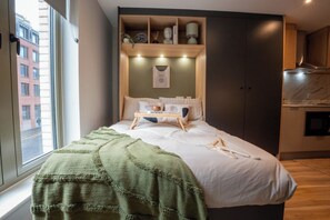 Ein Schlafzimmer mit einem Bett, einer grünen Decke, einem hölzernen Regal über dem Bett und Blick auf die Straße durch ein Fenster.