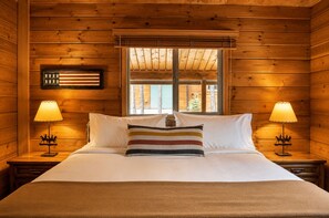 A wooden cabin room with a bed, two nightstands with lamps, a window with a view, and a flag on the wall. - Black Butte Ranch South Meadow 177 (Black Butte Ranch)