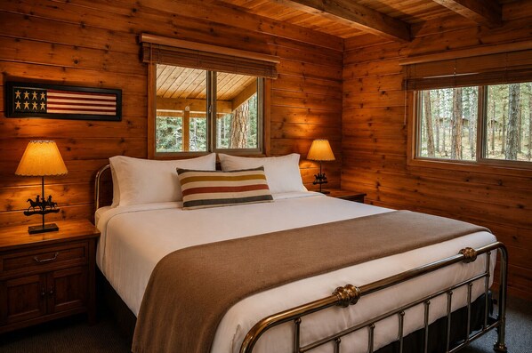 A wooden cabin bedroom with a bed, nightstands, lamps, a framed American flag, and a window with a view of trees. - Black Butte Ranch South Meadow 177 (Black Butte Ranch)
