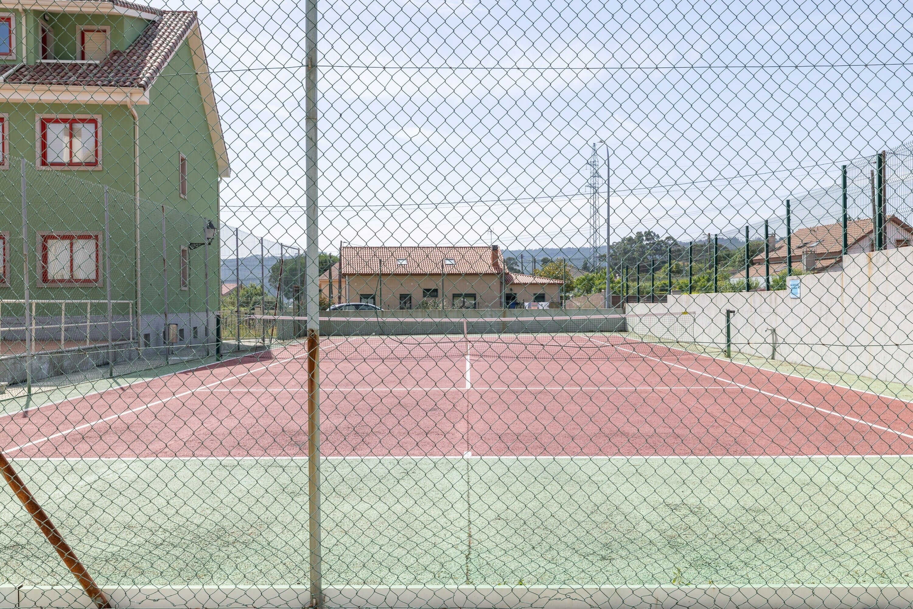 Un court de tennis entouré d’une clôture en treillis métallique, un bâtiment aux fenêtres rouges et un ciel dégagé.