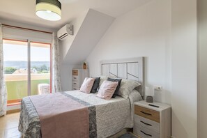 Um quarto com cama, mesas de cabeceira, abajur, janela com cortinas e aparelho de ar condicionado.