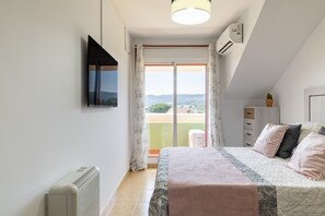 Un dormitorio con cama, televisión, radiador, ventana con cortinas y vistas al exterior.