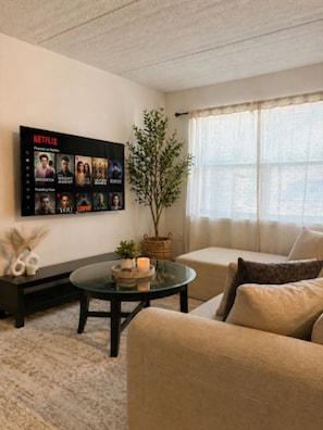 A living room with a flat-screen TV displaying a Netflix interface, a potted plant, a glass coffee table, and a beige sofa.