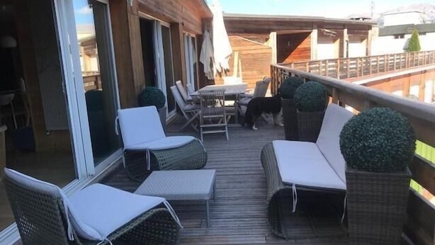 Una terraza de madera con sillas de mimbre y una mesa, una puerta de cristal que da a un espacio interior, y vistas al cielo.