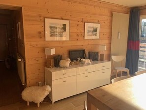 Ein Zimmer mit Holzwänden, einem weißen Schminktisch, zwei gerahmten Bildern an der Wand, einem Stuhl und einem kleinen weißen Hund.