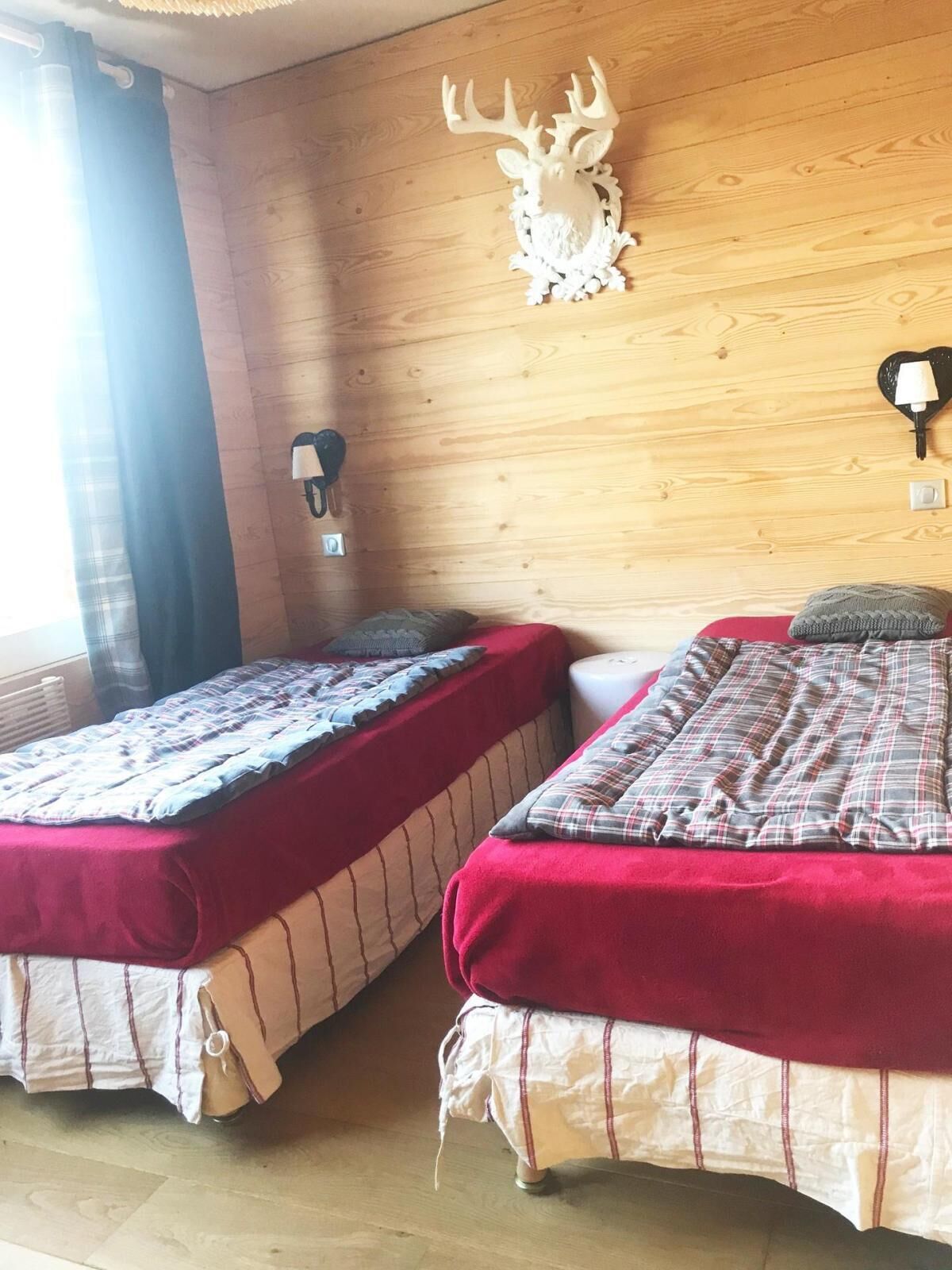 Two single beds with red and white bedding, a wooden wall with a deer head decoration, and a wall-mounted lamp.