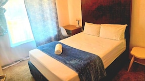 A hotel room with a bed, a nightstand, a chair, and a window with curtains. - Room-aquasaints Guest Lodge (Johannesburg)