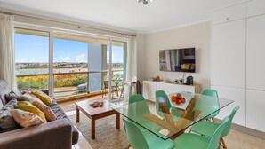 Moderni olohuone, jossa on lasinen ruokapöytä, vihreät tuolit, värikkäillä koristetyynyillä varustettu sohva ja suuri liukuovi, joka johtaa parvekkeelle, jolta avautuu näkymä kaupunkiin.