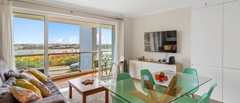 A modern living room with a glass dining table, green chairs, a sofa with colorful cushions, and a large sliding glass door leading to a balcony with a city view.
