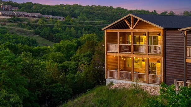 Eine Holzblockhütte mit mehreren Balkonen, umgeben von üppiger Vegetation und einer hügeligen Landschaft.