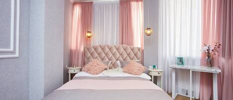Un dormitorio con cabecera capitonada, cama con ropa de cama rosa, dos mesitas de noche y una mesilla.