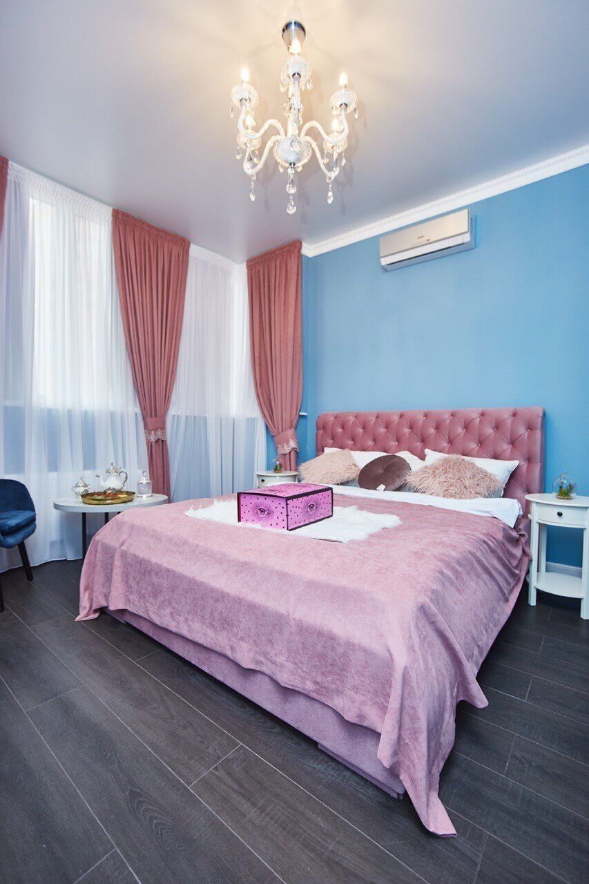 A bedroom with a pink bedspread, a chandelier, a bedside table, and a blue wall.