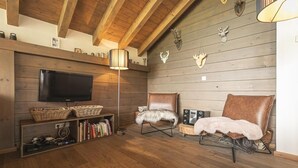 A cozy living room with a TV, bookshelves, two leather chairs, and wooden walls.
