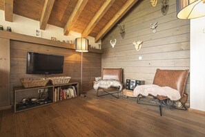 A cozy living room with a TV, bookshelves, two leather chairs, and wooden walls.