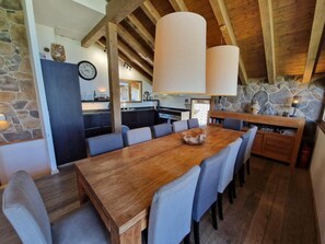 A dining area with a wooden table and chairs, a stone wall, and a kitchen in the background.
