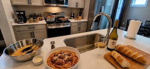 Une cuisine équipée d’une cuisinière, d’un évier et de divers ustensiles de cuisine. On y trouve un saladier de spaghettis, une bouteille de vin et un pain sur le comptoir.