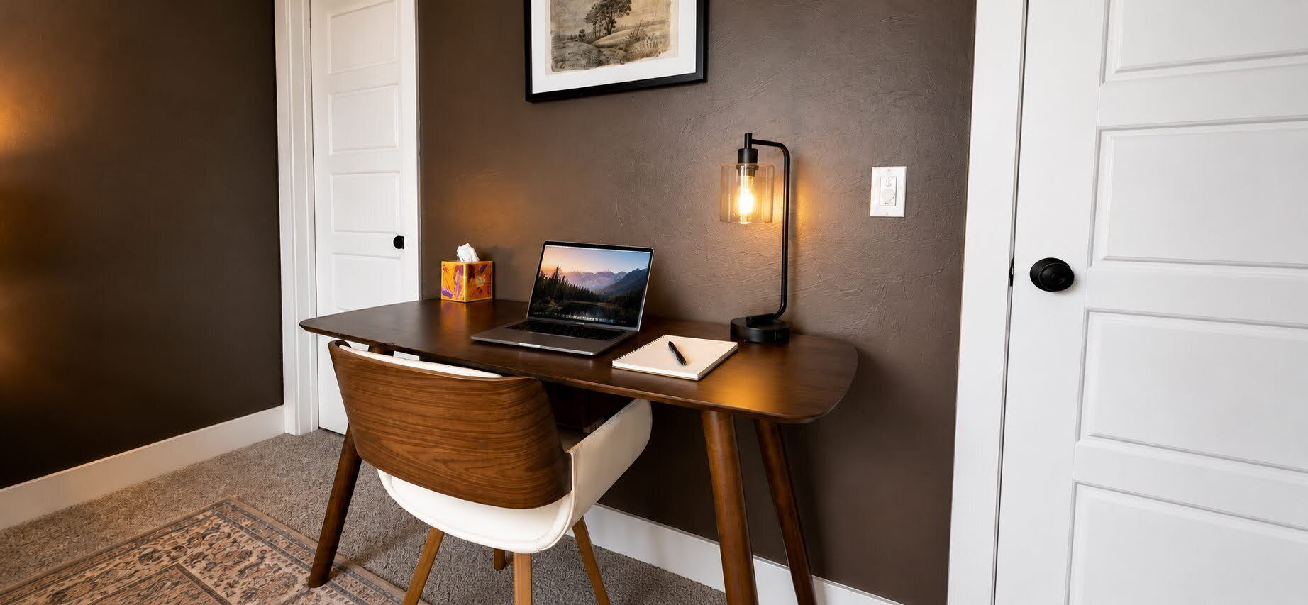 Een bureau met een laptop, een notitieboekje, een pen, een stoel, een zakdoos en een wandlamp.