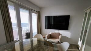 A modern room with a glass table, white chairs, a beige sofa, and a mounted TV. - für 2 Personen  + 4 Kinder ca. 52 m² in Norderney, Norddeutschland (Nieder by Interhome (Norderney)