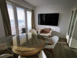 Ein modernes Zimmer mit Glastisch, weißen Stühlen, einem beigen Sofa und einem Wand-Fernseher.