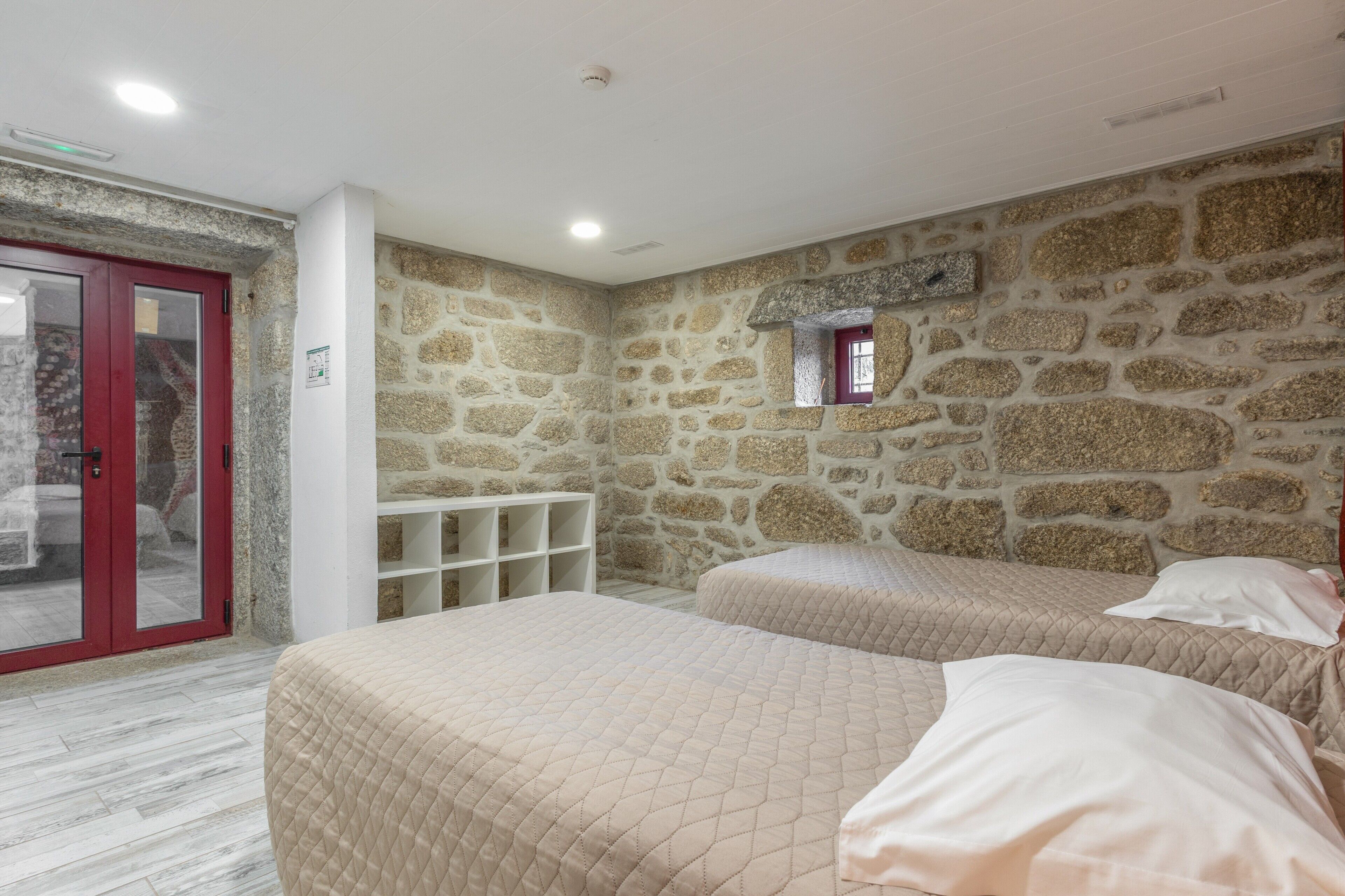 Un dormitorio con paredes de piedra, dos camas, una puerta roja y una ventana pequeña.