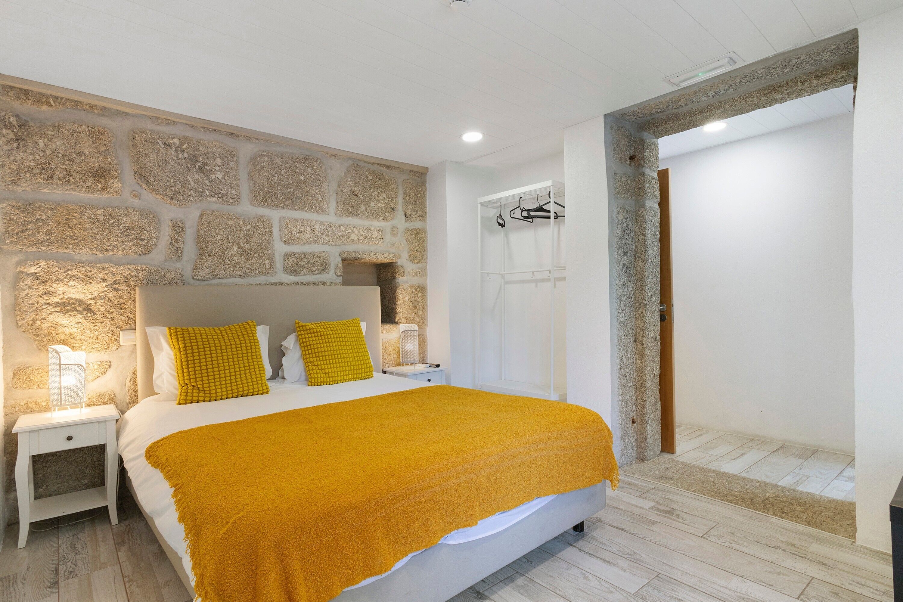 A bedroom with a stone wall, a bed with a yellow blanket, a nightstand, a white wardrobe, and a door leading to a bathroom.