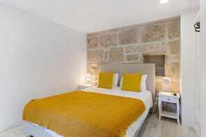 Un dormitori amb una paret de pedra, un llit amb llençols grocs, dues taules de nit amb làmpades i un armari blanc.