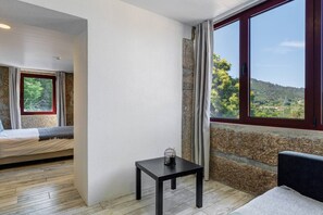 Une piĂšce avec un lit, une petite table, une fenĂȘtre donnant sur un paysage verdoyant et un mur en pierre.