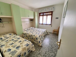 Ein Zimmer mit zwei Betten, einem kleinen Tisch, einem Fenster mit Vorhängen und einer Tür.