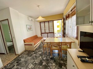 Ein kompakter Wohnbereich mit einer Couch, Esstisch, Stühlen, einem Fernseher und einem Fenster mit Jalousien.
