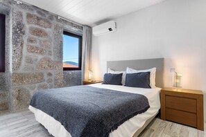 Une chambre avec un mur en pierre, un lit recouvert d’une couverture grise, une table de chevet avec une lampe et un appareil de climatisation.