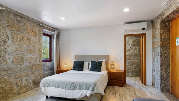 Un dormitorio con pared de piedra, una cama con colcha blanco y gris, dos mesitas de noche con lámparas y un equipo de aire acondicionado en la pared.