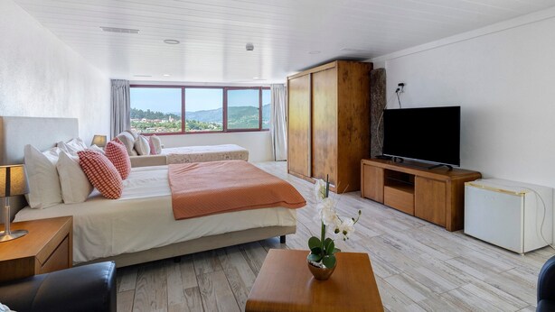 Ein modernes Hotelzimmer mit einem großen Bett, einem Fernseher, einer Holzkommode und Blick ins Freie.