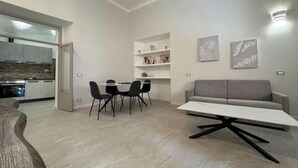 A modern living room with a sofa, a dining table with chairs, and a kitchen area visible through an open door.