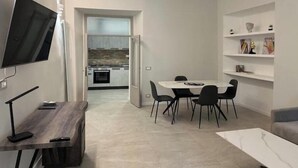 A modern living room with a dining area, a TV mounted on the wall, and a kitchen visible through an open door.