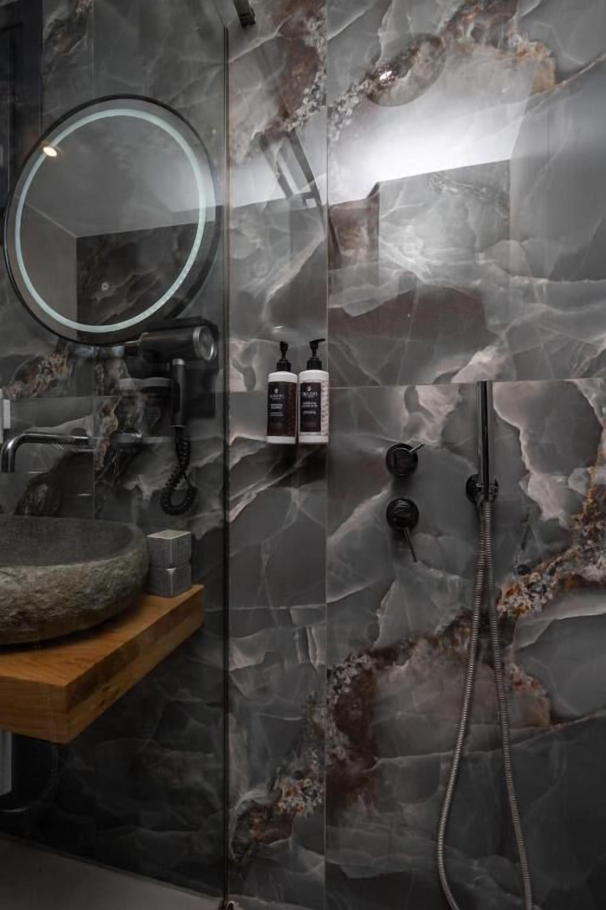 Une salle de bain avec un lavabo en pierre, un miroir rond, une douchette à main et une cabine de douche en verre.