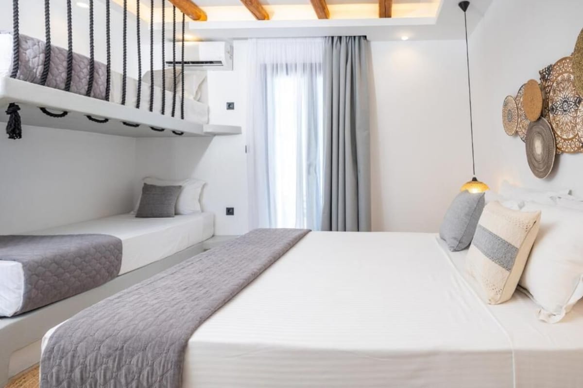 Quarto com cama, duas mesas de cabeceira, janela com cortinas, luminária pendente e um objeto decorativo fixado na parede.
