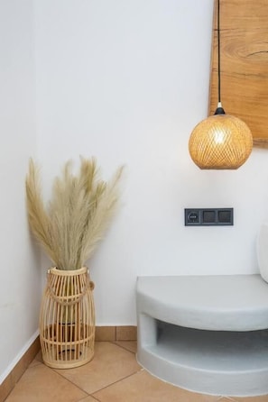 Un coin de pièce avec un mur blanc, un plancher en bois, un vase en osier contenant de l’herbe des pampas, un banc gris et un luminaire suspendu en osier.