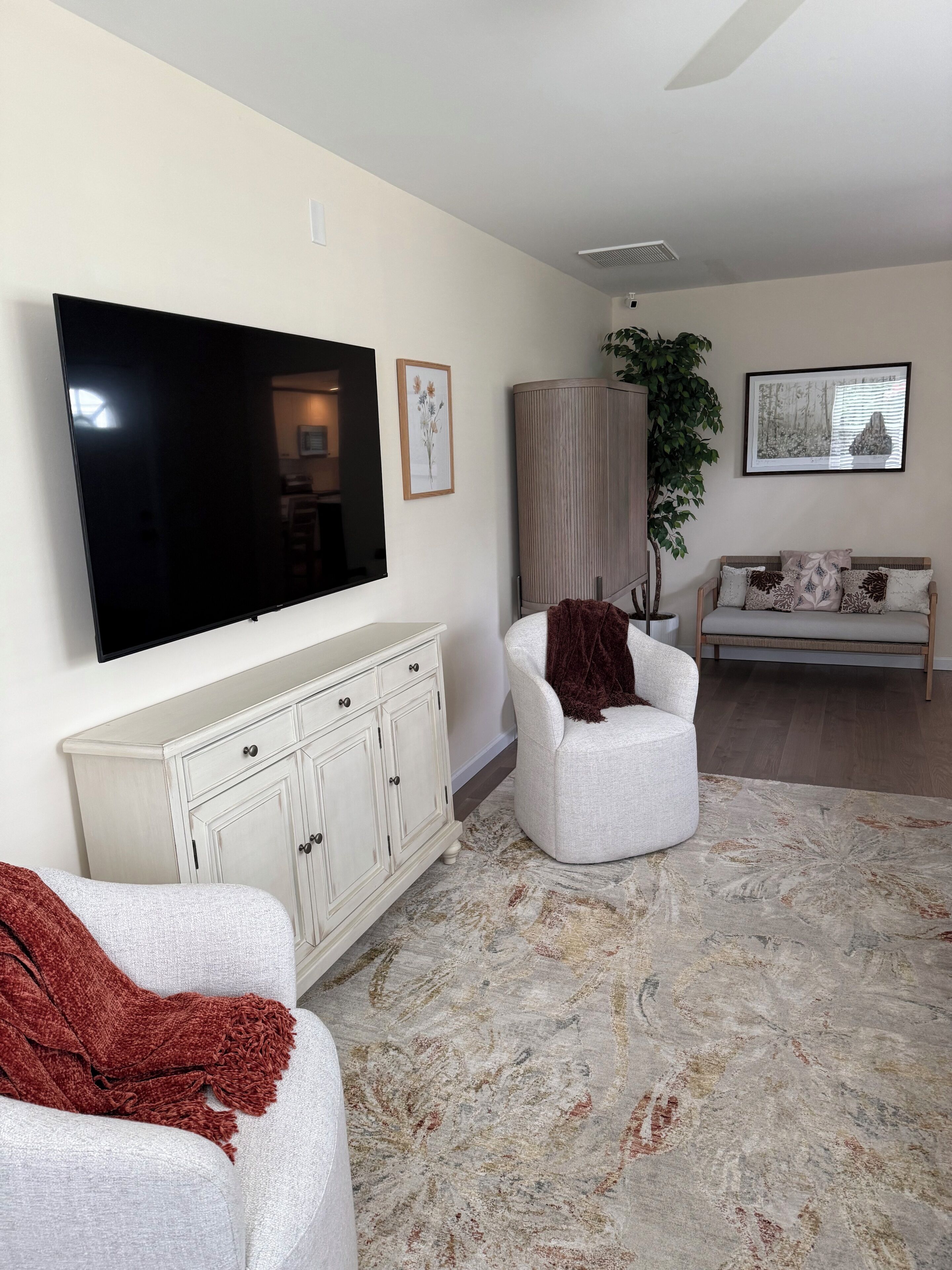 Un salon équipé d’un téléviseur à écran plat, d’un canapé blanc, d’un fauteuil blanc, d’un tapis à motifs et d’un meuble.