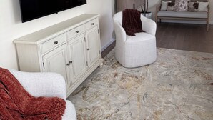 Un salon équipé d’un téléviseur à écran plat, d’un canapé blanc, d’un fauteuil blanc, d’un tapis à motifs et d’un meuble.