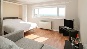 A bedroom with a bed, a TV, a window with a beach view, and a small table with glasses. - Flat 30 (Bridport)