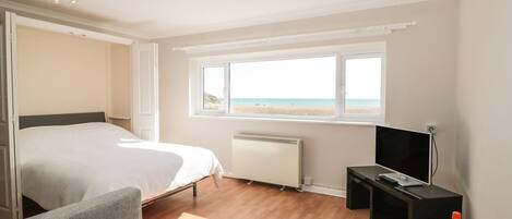 A bedroom with a bed, a TV, a window with a beach view, and a small table with glasses.
