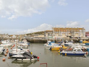 A marina with various boats docked, a waterfront building, and a hilly landscape in the background. - Flat 30 (Bridport)