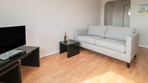 A living room with a flat-screen TV, a grey sofa, and a small black side table with a bottle and glass. - Flat 30 (Bridport)