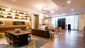 Eine geräumige Hotellobby mit einem Holzbüro, einer Sitzecke mit Sofas, einem Bücherregal voller Bücher und einem großen Fenster, das natürliches Licht hereinlässt.