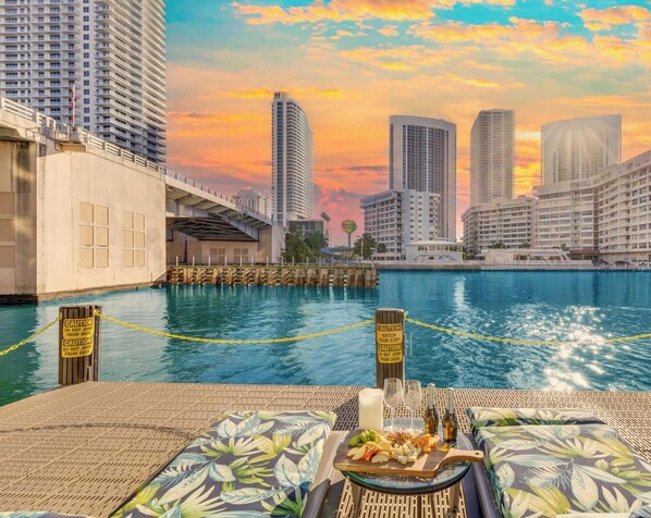 A waterfront view with high-rise buildings, a dock, and a dining setup with a plate of food and drinks. - Chic 1BR Steps from the Beach Shuttle (Hallandale Beach)