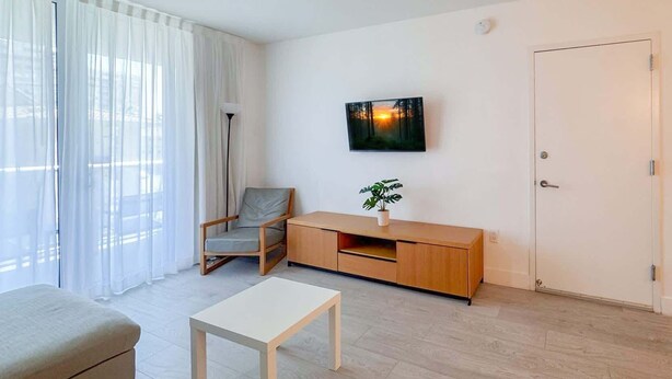 Ett modernt vardagsrum med en soffa, ett soffbord, en fåtölj, en tv på väggen, en växt på ett träskåp och en dörr.