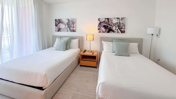 A hotel room with two beds, a nightstand with a lamp, and artwork on the walls. - Beachwalk Studio | Intracoastal View (Hallandale Beach)
