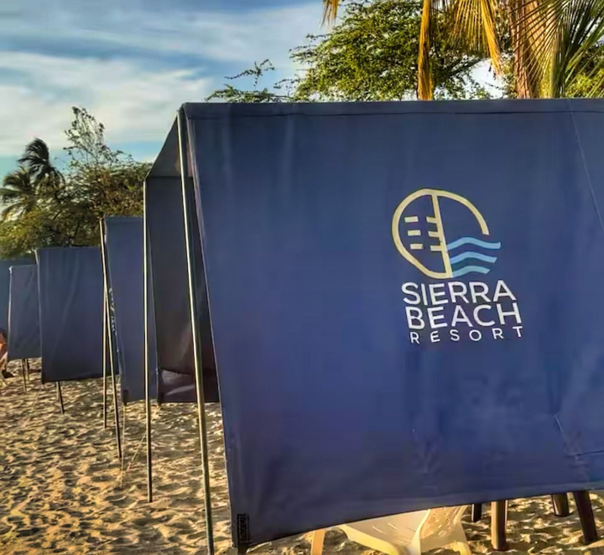 Una fila de carpas con el logotipo de Sierra Beach Resort, sobre una playa arenosa con palmeras al fondo.
