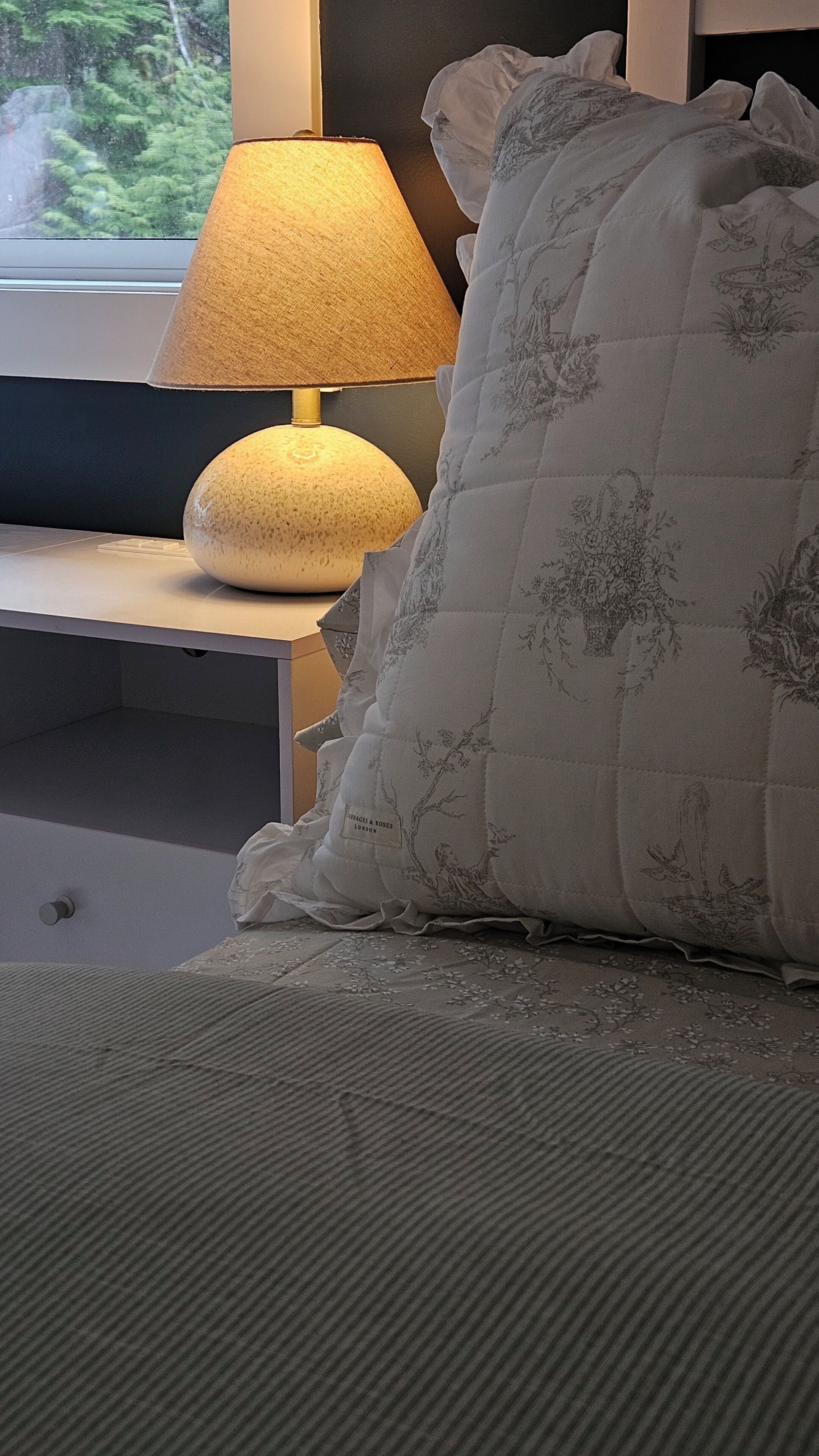 Un lit recouvert d’une couverture grise à texture marquée, un oreiller blanc orné de broderies florales et une table de chevet avec une lampe.