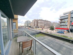 Ein Balkon mit Tisch und Stuhl, der auf eine Straße mit Gebäuden und einem Gebäude mit rotem Dach blickt.